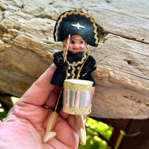 May include: A vintage Christmas ornament of a drummer boy. The doll is dressed in a black uniform with gold trim and a matching hat. The drum is silver with gold accents, and the doll is holding drumsticks. The ornament is approximately 4 inches tall.