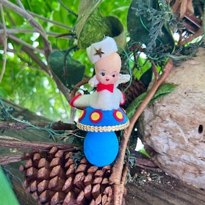 Może przedstawiać: Zabytkowa ozdoba choinkowa przedstawiająca postać przypominającą lalkę z białym kapeluszem i gwiazdą, czerwoną muszką i niebieską podstawą w kształcie grzyba. Ozdoba ma niebieski, czerwony i złoty wzór. Wisi wśród zielonych liści i gałęzi.