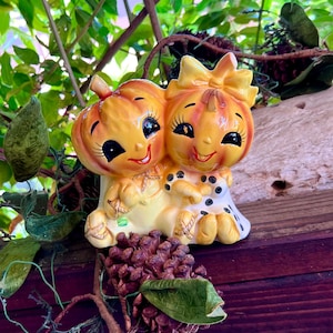 May include: A ceramic figurine featuring two anthropomorphic pumpkin characters with smiling faces, large eyes, and long eyelashes. One pumpkin wears a yellow bow, while the other has a stem top. They are joined together in a seated position.