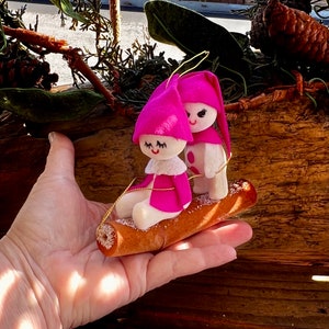 May include: A vintage Christmas ornament featuring two elf figures with pink hats and coats, seated on a brown log. The elves have white faces and are connected by a gold string. The ornament is held in a hand.