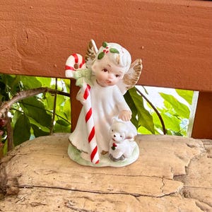 May include: A ceramic figurine of an angel with gold-painted wings, wearing a white robe, holding a red and white striped candy cane, and standing next to a small white lamb. The angel has holly leaves with red berries in its hair.
