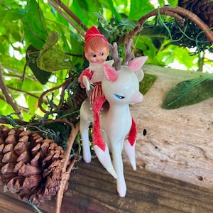 Op de afbeelding: Een vintage kerstornament met een wit hert met roze oren en blauwe ogen, bereden door een pop in een rode hoed en outfit. Het hert heeft rode accenten. Het ornament staat tegen een natuurlijke achtergrond met groen en een dennenappel.