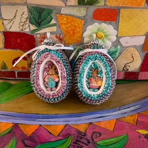 以下が含まれることがあります： ピンクとティール色のビーズで装飾された2つのイースターエッグオーナメント。それぞれの中に小さなウサギの置物が入っています。オーナメントはスパンコールとリボンで飾られ、カラフルなモザイクの背景に配置されています。