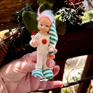 May include: A small doll with a pale face, blue eyes, and pink hair, wearing a white ribbed jumpsuit with a round orange patch on the chest. The doll has a green and white striped nightcap and matching slippers, and holds a small white candle.