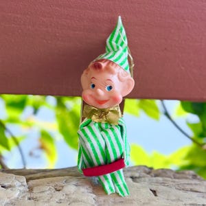 Puede incluir: Un adorno de elfo de estilo vintage con un gorro puntiagudo y ropa de tela blanca con rayas verdes. El elfo tiene una cara de plástico moldeado con ojos azules, una pequeña sonrisa, una pajarita dorada y una banda roja sobre las piernas.