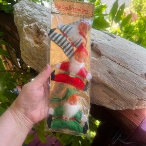 May include: A package of three vintage-style holiday ornaments featuring figures with plastic faces and white beards. The top figure wears green and white striped clothing, the middle wears red, and the bottom wears green. The header reads Holiday Decorations.
