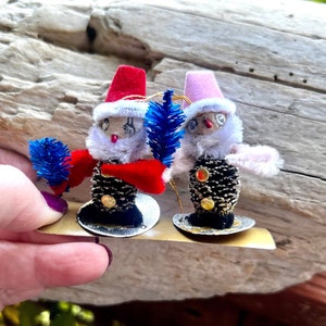 May include: Two vintage Christmas ornaments shaped like snowmen. Each snowman has a pinecone body, a red or pink hat, and a fluffy white beard. They hold blue pipe cleaner trees and are mounted on a gold-colored base.
