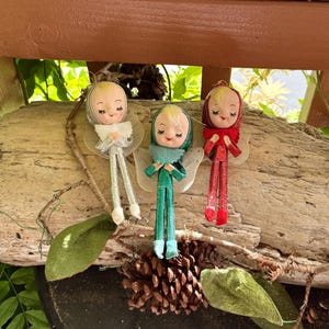 Könnte beinhalten: Drei Wichtel-Ornamente im Vintage-Stil mit bemalten Gesichtern, geschlossenen Augen und rosigen Wangen. Jeder Wichtel trägt eine spitze Kapuze und ein passendes Outfit aus silbernem, grünem oder rotem Glitzerstoff mit Rüschenkragen und Flügeln.