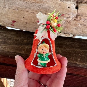 May include: A vintage Christmas bell ornament. The bell is red with a silver glitter trim around the opening. Inside the bell is a small figurine of a child wearing a green outfit. The top of the bell is decorated with white paper flowers, red berries, and green sprigs.