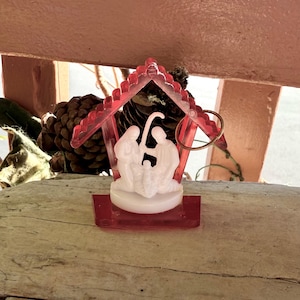 Può includere: Un piccolo presepe in plastica rossa con una statuina bianca di Maria, Giuseppe e Gesù bambino. La scena è racchiusa in un rifugio di plastica rossa con un anello dorato attaccato. Una pigna è visibile sullo sfondo.