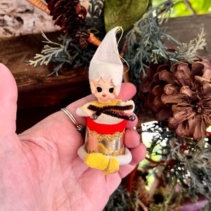 May include: A vintage-style elf ornament featuring a white felt hat, a yellow shirt, and a red and gold drum. The elf has painted facial features, pointed ears, and holds two brown pipe cleaner drumsticks with small silver-colored tips.
