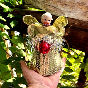 May include: A vintage Christmas angel ornament. It features a gold mesh skirt, gold foil wings, and a small porcelain face. The angel has a red bauble and silver tinsel. A festive holiday decoration.