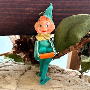 May include: A vintage elf figurine with a painted face, green felt outfit, and a gold bow tie. The elf is holding an orange object and has a green hat. The figurine is placed on a wooden surface with a leaf and pinecone.