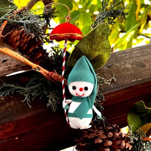 May include: A vintage Christmas ornament featuring a snowman figure with a red parasol. The snowman has a white body, green hat and scarf, and a smiling face. The parasol is red with gold trim and a striped pole. The ornament is hanging among pinecones and greenery.