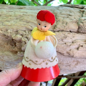 May include: A decorative figurine featuring a doll with a red felt cap, a yellow felt collar, and a white felt dress with gold glitter trim. The doll has a painted face and holds a white spherical object in its hands, with a solid red base.