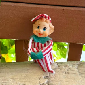 Puede incluir: Una pequeña figura de elfo con cara de plástico moldeado, ojos azules y expresión sonriente. El elfo viste un traje de tela a rayas rojas y blancas con un gorro puntiagudo a juego, complementado con un cuello y manoplas de fieltro de color verde.