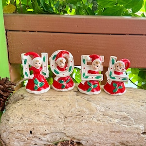 May include: Four ceramic figurines spelling out the word NOEL. Each figurine features a child wearing a red Santa hat and dress, accented with green holly leaves and red berries. The letters are white with green leaf details and red trim.
