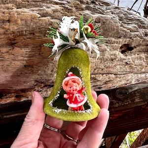 Può includere: Un ornamento a campana natalizia vintage. La campana è di colore verde oliva sfocato con un ritaglio che rivela una piccola statuina in un abito rosso e bianco. La parte superiore della campana è decorata con fiori bianchi, bacche rosse e accenti dorati.