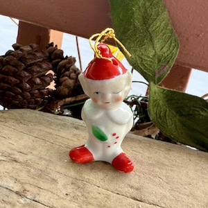 May include: A vintage ceramic Christmas elf figurine. The elf is wearing a white outfit with red shoes and a red hat. It has a green leaf and red dots on its chest. A gold string is attached to the hat for hanging.