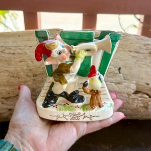 Könnte beinhalten: Eine Vintage-Keramikfigur eines skurrilen Elfen, der Trompete spielt, mit einer kleineren Vogelfigur. Der Elf trägt einen roten Hut, eine braune Jacke und schwarze Stiefel. Der Hintergrund hat grüne und weiße Streifen. Die Figur wird in einer Hand gehalten.