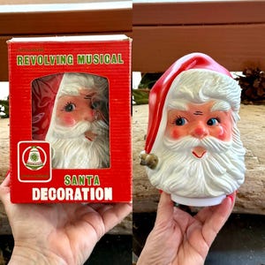 May include: A revolving musical Santa decoration featuring a molded plastic head with a red hat, white beard, and rosy cheeks. The item is packaged in a red cardboard box with the text REVOLVING MUSICAL SANTA DECORATION and a bell logo.