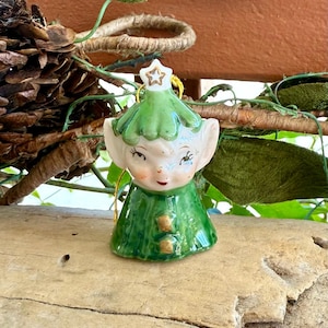 May include: A ceramic Christmas ornament in the shape of a winking elf. The elf has a green hat with a gold star, a green tunic, and rosy cheeks. The ornament is on a wooden surface.
