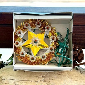 May include: A gold tinsel Christmas tree topper featuring a central five-pointed star with a reflective finish. The star is surrounded by a circular frame adorned with ten small, clear plastic light covers, each containing a miniature bulb.