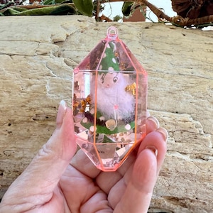 May include: A pink, geometric-shaped ornament with a clear center, showcasing a fluffy white figure and green accents. Gold-colored embellishments are visible within the ornament. The ornament is held by a hand.