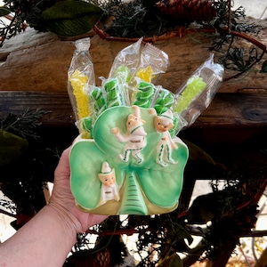 Könnte beinhalten: Ein grüner Kleeblatt-förmiger Keramikbehälter mit drei Elfenfiguren, gefüllt mit verpackten Süßigkeiten. Die Süßigkeiten sind gelb und grün, und der Behälter wird von einer Hand gehalten.