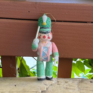 Könnte beinhalten: Ein glitzerndes Spielzeugsoldaten-Ornament mit grünem Hut und weißer Feder, rosa Jacke und grüner Hose. Die Figur hält einen weißen Trommelstock und steht hinter einer kleinen blau-rosa Trommel, inklusive einer goldenen Aufhängeschnur.