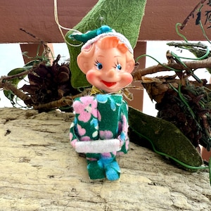 Peut inclure: Une figurine d'elfe vintage avec un visage en plastique, des yeux bleus peints et une bouche rouge. L'elfe porte un chapeau en tissu vert et une tenue assortie à motif floral rose et bleu, tenant un petit objet rectangulaire blanc dans ses mains.