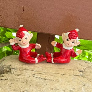 May include: Two ceramic elf figurines in a seated position, each wearing a red suit with white trim and a matching red pointed hat. The elves have painted facial features, including dark eyes and eyebrows, and are posed with arms extended.