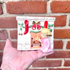 May include: A white ceramic Christmas fireplace with red stockings, gold bells, and a small figure in a pink dress. The fireplace has a textured surface and a painted fire inside.