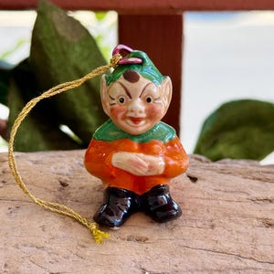 Peut inclure: Figurine d'elfe en céramique avec un chapeau et un col verts, une chemise orange et des bottes noires. L'elfe a un visage souriant avec des joues roses et une ficelle dorée pour l'accrocher. La figurine est sur un morceau de bois.