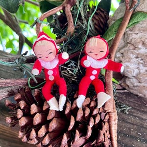 Op de afbeelding: Twee kleine elf-ornamenten gemaakt van rood vilt met witte randen op de mutsen, kragen en voeten. Elke elf heeft een geschilderd gezicht met blond haar, roze wangen en een glimlach. De figuren hangen aan dunne witte koordjes.