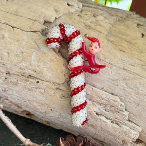 Puede incluir: Un adorno de bastón de caramelo decorado con rayas diagonales alternas de lentejuelas rojas y blancas iridiscentes. Una pequeña figura de elfo roja brillante con un sombrero puntiagudo y rasgos faciales pintados está unida al bastón.