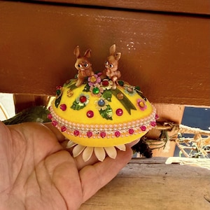 以下が含まれることがあります： 2匹の小さなウサギの置物が乗った、装飾的なイースターエッグ。卵は黄色で、カラフルな花、宝石、真珠の縁取りで飾られています。ベースには白い花びらのようなアクセントがあります。