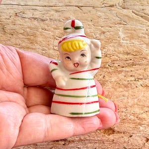 Puede incluir: Pequeña figura de cerámica vintage de un niño con un atuendo a rayas con detalles en rojo y verde. La figura tiene una cara sonriente, cabello amarillo y un sombrero a juego. Sostenido en una mano.