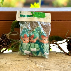 May include: A package of three vintage pixie ornaments with green felt outfits and pointed hats. The header card reads Dexter's Green Holly Pixies, Catalog No. 20-17168, Made in JAPAN. Each pixie has a painted face with rosy cheeks and red hair.