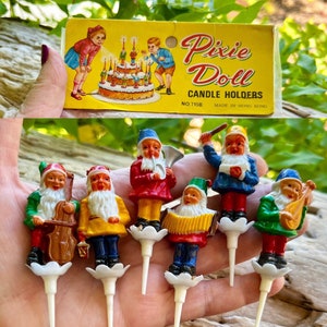 Peut inclure: Ensemble de sept bougeoirs en plastique en forme de gnomes colorés. Chaque gnome joue d'un instrument de musique différent. L'emballage montre un gâteau d'anniversaire avec des bougies et le texte "Pixie Doll Candle Holders No. 715B Made in Hong Kong."