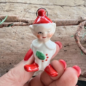 May include: A vintage Christmas elf ornament. The ceramic figurine features a red hat and boots, a white collar, and a winking face. The elf is holding a green leaf and red berries. A small wire loop is attached for hanging.