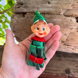Pode incluir: Um ornamento de elfo em estilo vintage com rosto de plástico moldado, olhos azuis pintados e lábios vermelhos. O elfo usa um chapéu de tecido verde metálico e uma túnica verde a condizer com um colarinho vermelho e um acessório retangular vermelho.