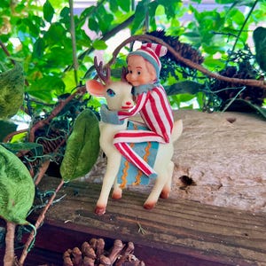 May include: A vintage Christmas figurine of a small elf in a red and white striped outfit riding a white reindeer with brown antlers. The reindeer has a blue and gold blanket. The figurine is set against a backdrop of green foliage and a wooden surface.