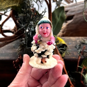 May include: A vintage Christmas ornament featuring a whimsical elf figure. The elf has a pink face, blue eyes, and a white hooded hat. The body is made of a pine cone, and it is adorned with pink trim and small bells. The ornament is on a round, cream-colored base.