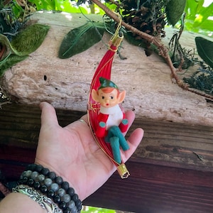 May include: A vintage Christmas ornament featuring a seated elf with red and green clothing, a green hat, and rosy cheeks. The elf is seated on a red, curved, woven base with gold accents. The ornament is suspended by a gold wire.
