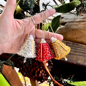 Op de afbeelding: Drie belvormige kerstversieringen in crème, rood en goud. Elk ornament is versierd met kralen en pailletten en heeft een goudkleurig ophangkoord. Een witte kraal en een bungelende parel accentueren elke bel.