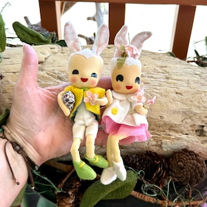 以下が含まれることがあります： 白いフェルトの耳と顔をした、ヴィンテージのイースターバニー人形2体。男の子のバニーは黄色のジャケット、白いズボン、緑色の靴を履いています。女の子のバニーは白いジャケットにピンクのスカート、白い靴を履いています。両方のバニーはピンクの頬をしていて、花を持っています。