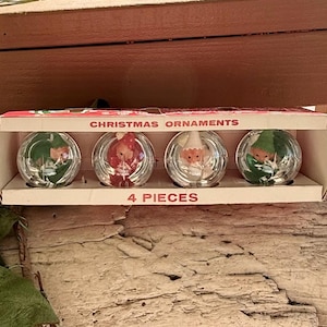 Puede incluir: Una caja de cuatro adornos navideños vintage. Cada adorno de cristal transparente presenta un diseño festivo diferente, incluyendo elfos y Papá Noel. La caja está etiquetada como "Christmas Ornaments" y "4 Pieces".