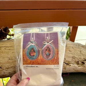 以下が含まれることがあります： 透明なビニール袋に入ったクラフトキット。キットには、それぞれウサギが入った2つのイースターエッグのデザインが印刷された紙が含まれています。紙には「Herrschners inc.」と「Easter Boutiques」と書かれています。