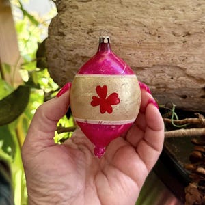 May include: A vintage Christmas ornament with a pink and gold color scheme. The ornament has a teardrop shape with a gold band featuring a red floral design. The top has a metal loop for hanging.